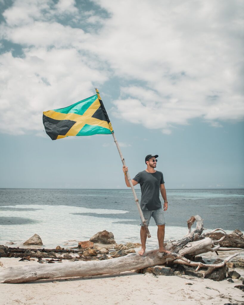 White man standing on a piece of drift wood on a beach in Negril waving a Jamaican flag and considering buying property in Jamaica